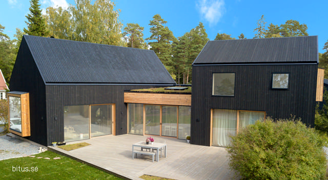 Ett modernt designat svart hus med stora fönsterpartier och ett stort trädäck.