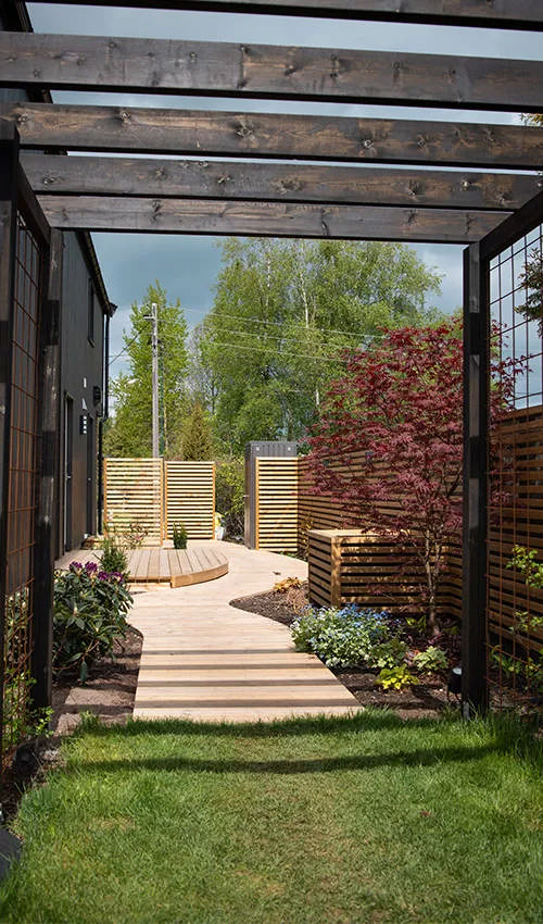 Ett trädäck i snirklig design vid ett modernt svart hus kantat av vackra planteringar.